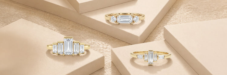 Dainty lab-grown diamond engagement rings in yellow gold — graduated baguette cluster ring, emerald cut three-stone with round brilliant side stones, and linear baguette bar solitaire on layered beige stone steps