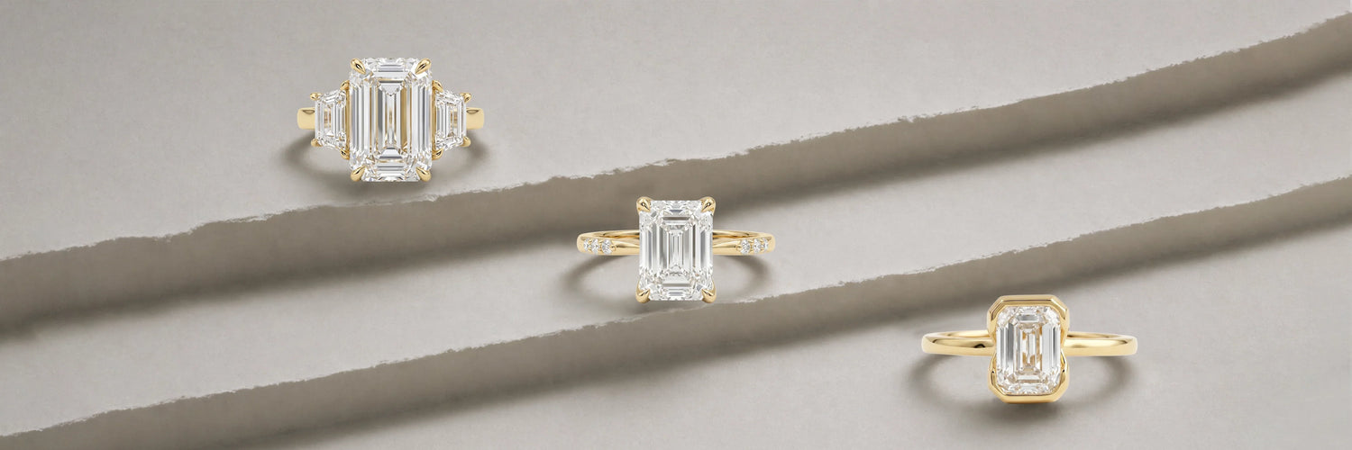 Three emerald cut lab-grown diamond engagement rings in yellow gold — three-stone with trapezoid baguette side stones, classic four-prong solitaire with thin band, and full bezel solitaire on layered stone surface