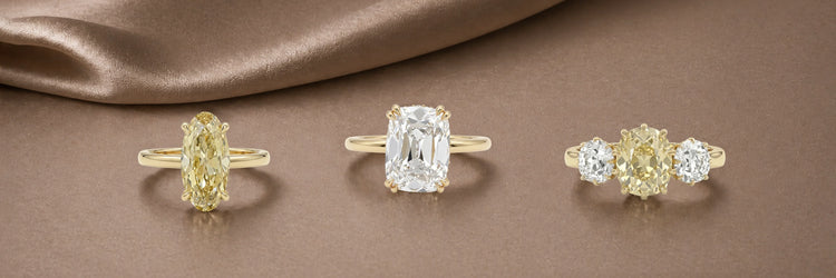 Three old cut lab-grown diamond engagement rings in yellow gold — fancy yellow oval solitaire, large old mine cushion cut solitaire with claw prongs, and fancy yellow three-stone ring with round white diamond side stones on champagne satin