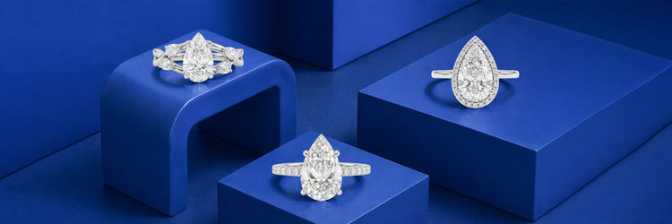 Three pear shaped lab-grown diamond engagement rings in white gold — vintage floral three-stone with marquise and baguette accents, large pear solitaire with pavé band, and pear cut diamond halo ring on cobalt blue display