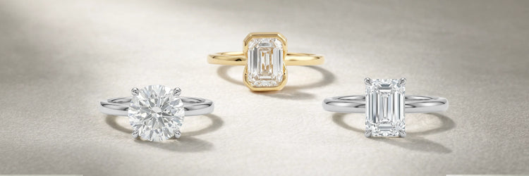 Three solitaire lab-grown diamond engagement rings displayed on a neutral surface — round brilliant cut in white gold, emerald cut bezel setting in yellow gold, and emerald cut four-prong solitaire in white gold
