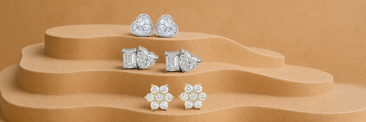 Lab-grown diamond stud earrings — heart cut halo studs in white gold, mixed emerald and heart cut two-stone studs in white gold, and round brilliant floral cluster studs in yellow gold on tiered camel sand display