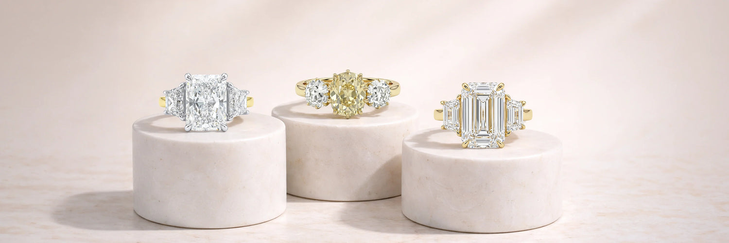 Three stone lab-grown diamond engagement rings displayed on white pedestals – radiant, oval yellow, and emerald cut in gold settings